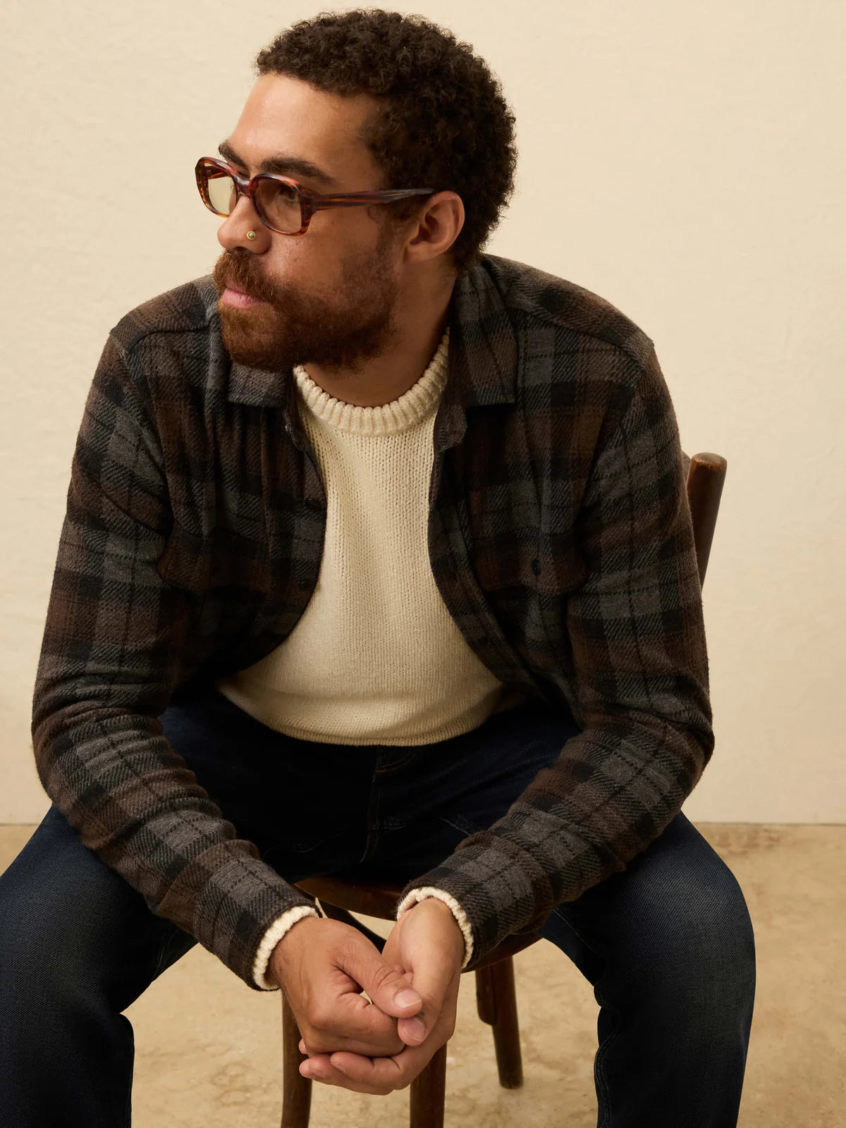 Man wearing glasses and a plaid shirt sitting on a chair against a beige background