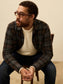 Man wearing glasses and a plaid shirt sitting on a chair against a beige background